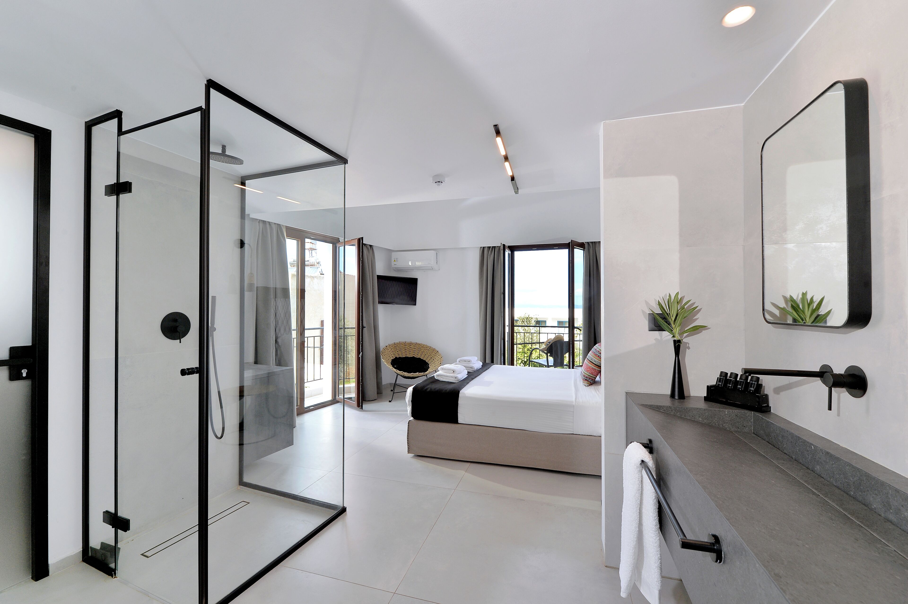 A modern hotel room with a glass-enclosed shower, a large bed, a bathroom with a vanity, and a balcony with a view.