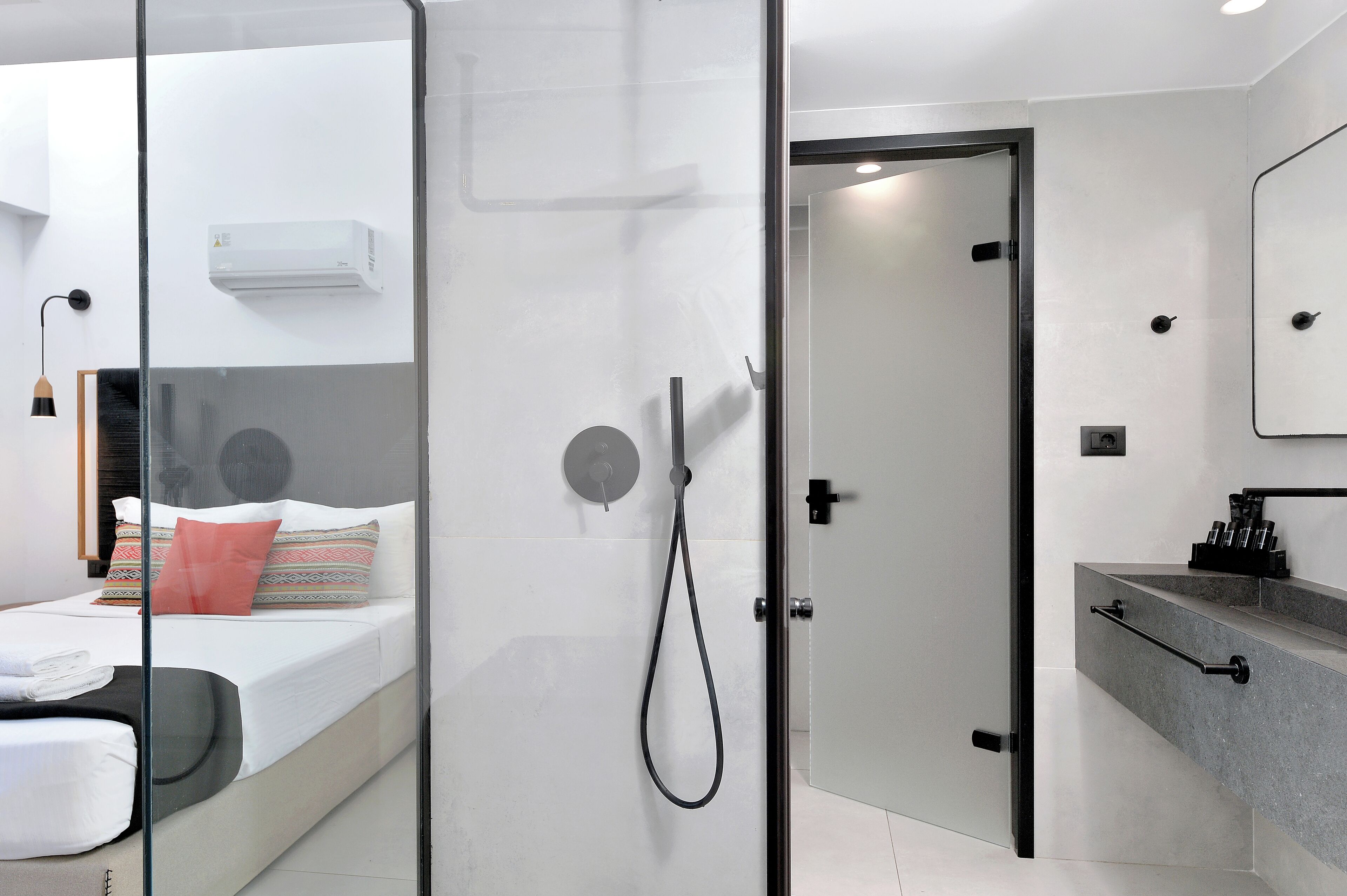 A modern hotel room with a glass shower enclosure, a bed with patterned pillows, a white air conditioner, and a bathroom with a grey countertop and a mirror.