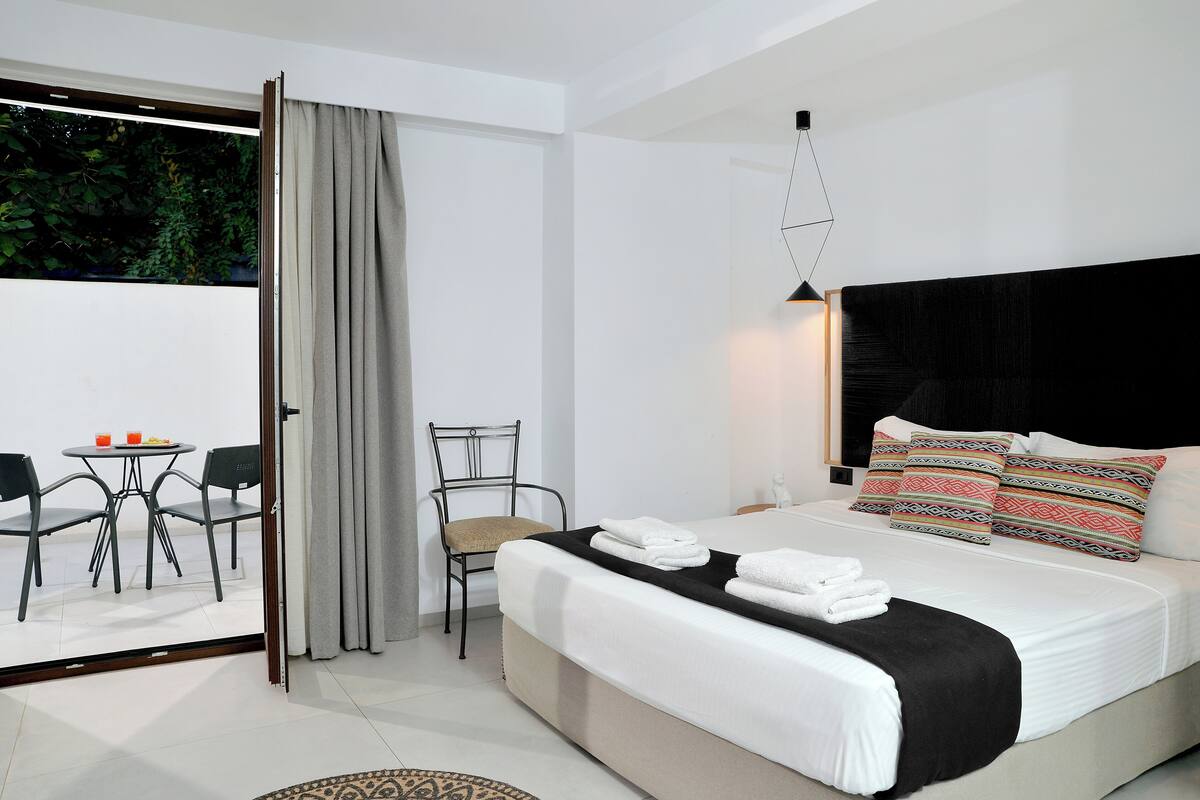 A modern hotel room with a large bed, a dining area with a table and chairs, and a view of greenery through a sliding glass door.