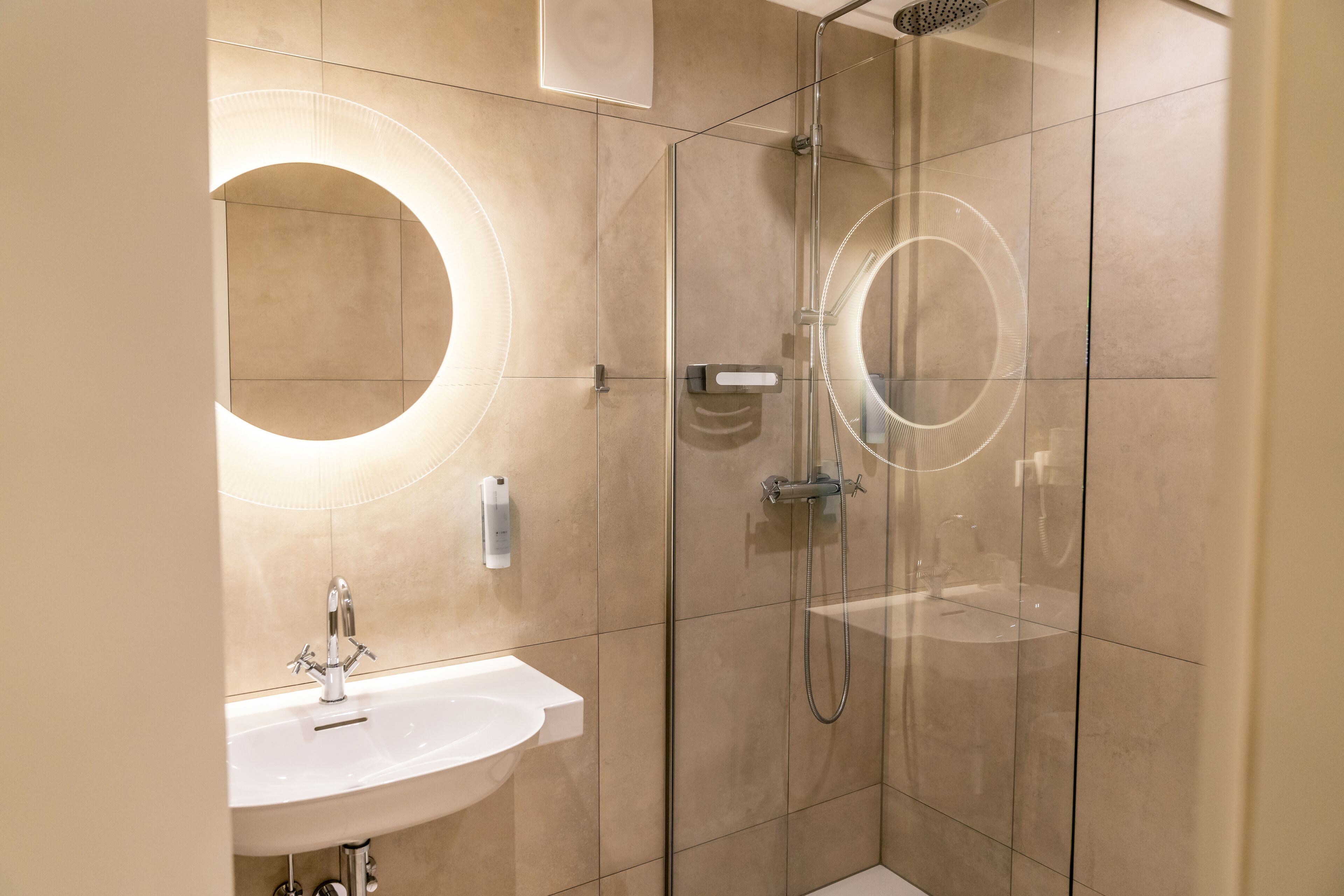 A modern bathroom with a glass-enclosed shower, a round mirror, and a white sink.