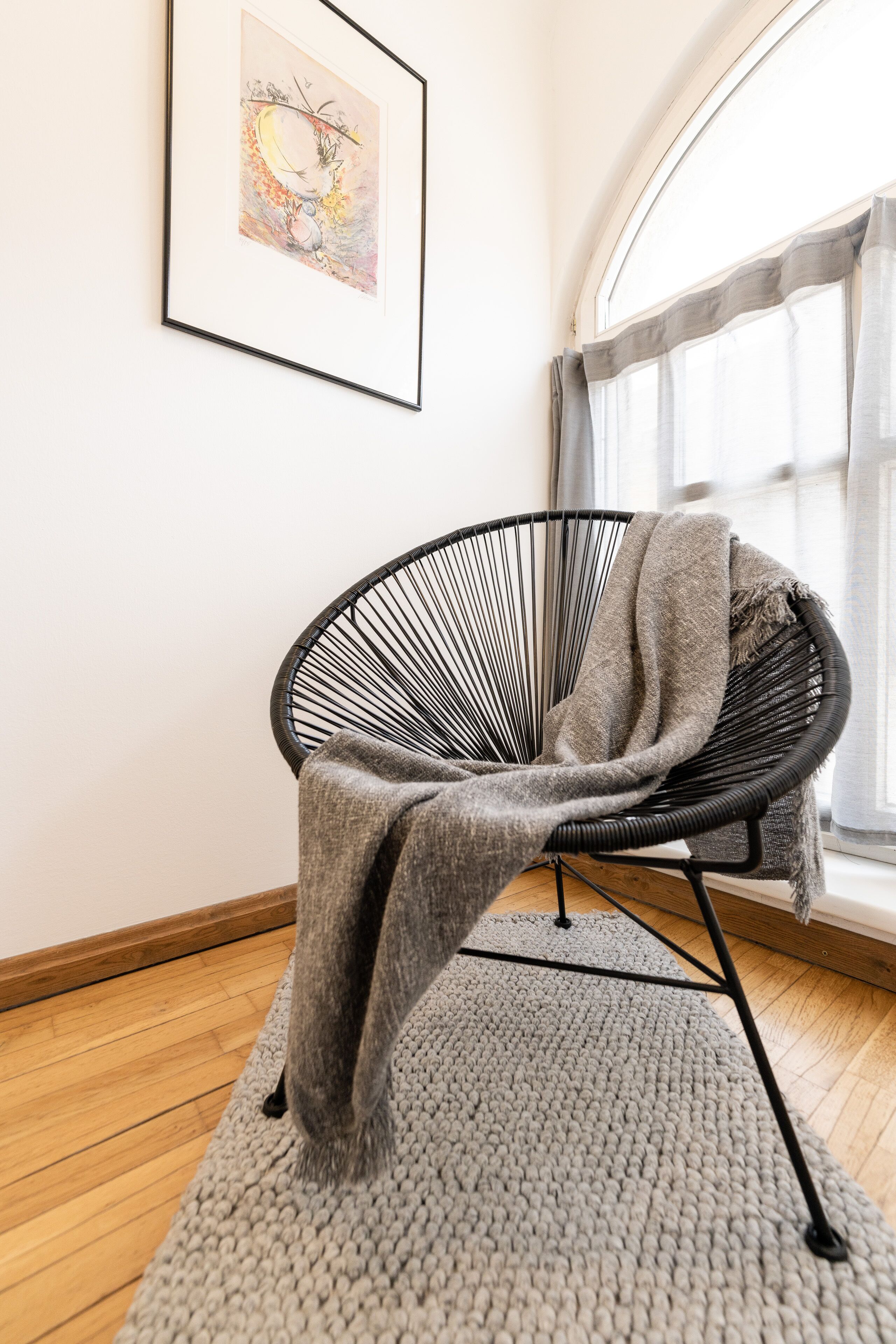 A modern room with a black wireframe chair, a grey knitted rug, and a framed artwork on the wall.