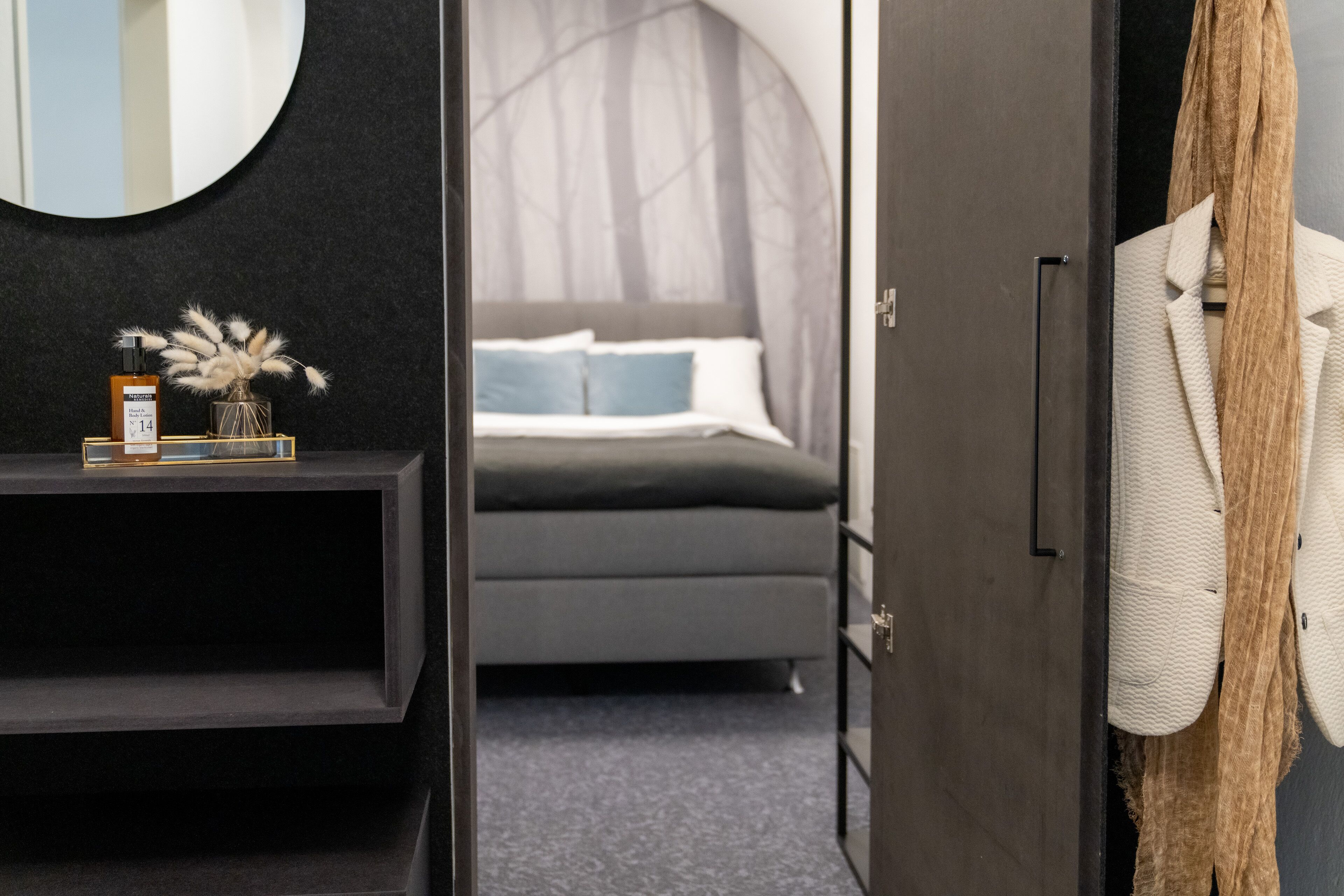 A bedroom with a bed, a bedside table with a bottle and a vase, and a coat rack with a coat.
