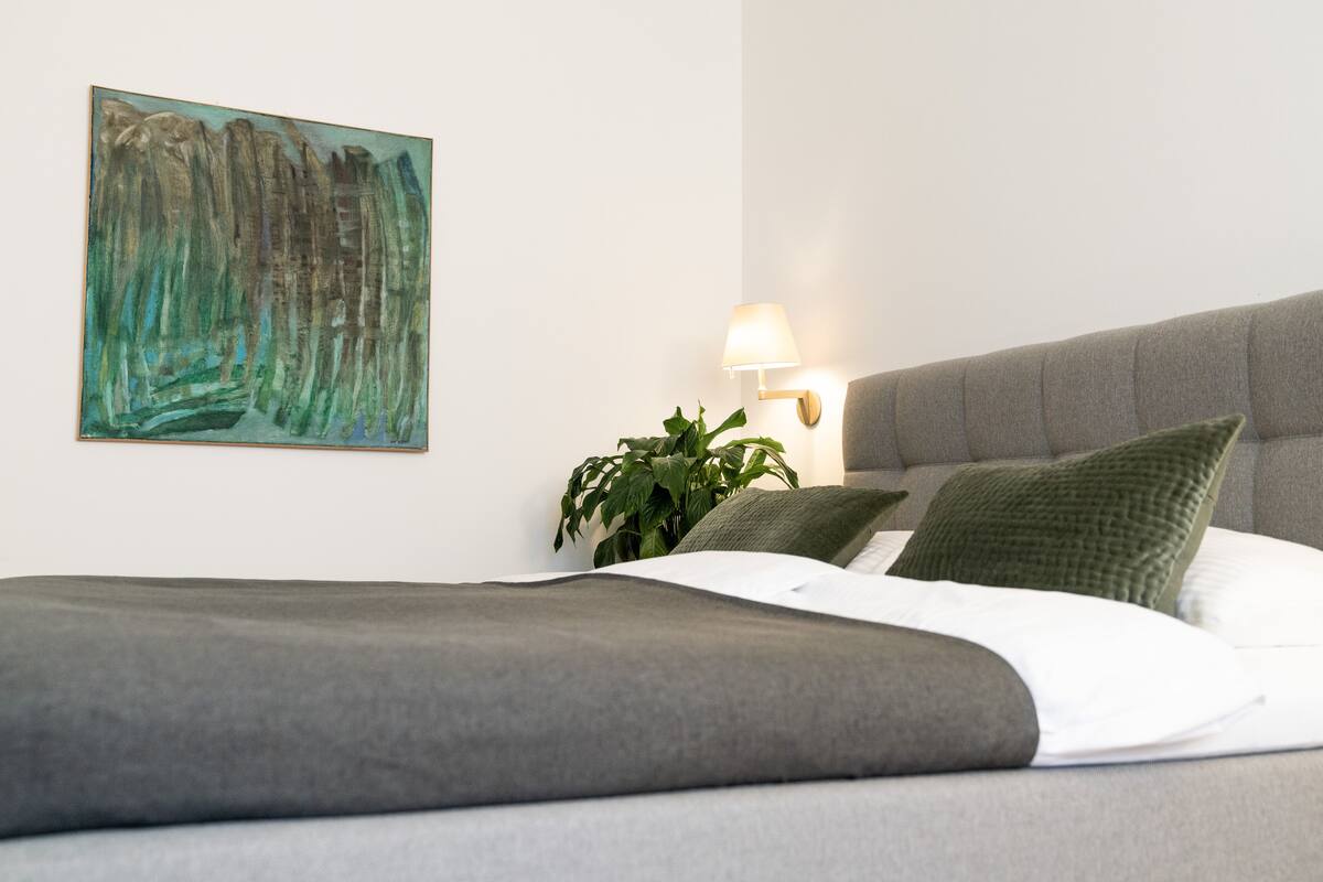 Una cama bien hecha con un cabecero gris, una almohada verde y una almohada blanca. Hay una lámpara de pared y un cuadro en la pared.