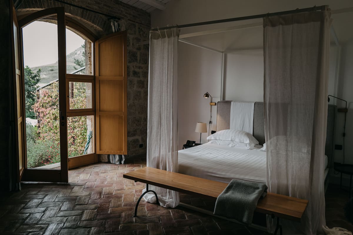 A bedroom with a four-poster bed, a wooden bench, a nightstand, and a view of greenery through an open window.