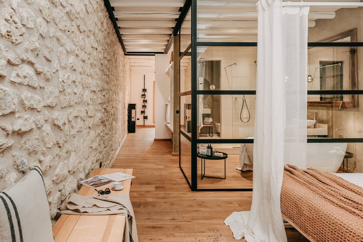 A modern hotel room with a wooden floor, a stone wall, and a glass partition. There is a bed with a white canopy, a small table with a newspaper and glasses, and a bathroom visible through the glass partition.