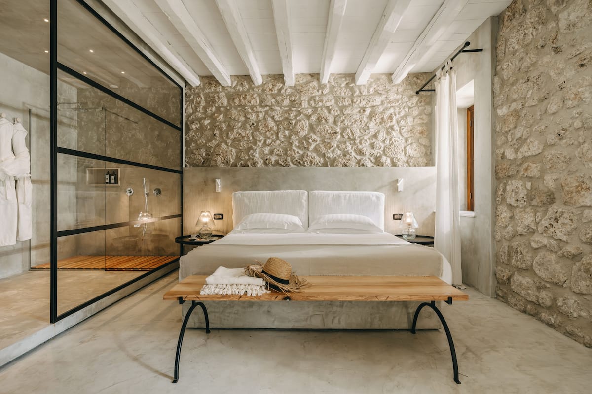 A modern bedroom with a stone wall, a large bed, a wooden bench, and a glass shower enclosure.
