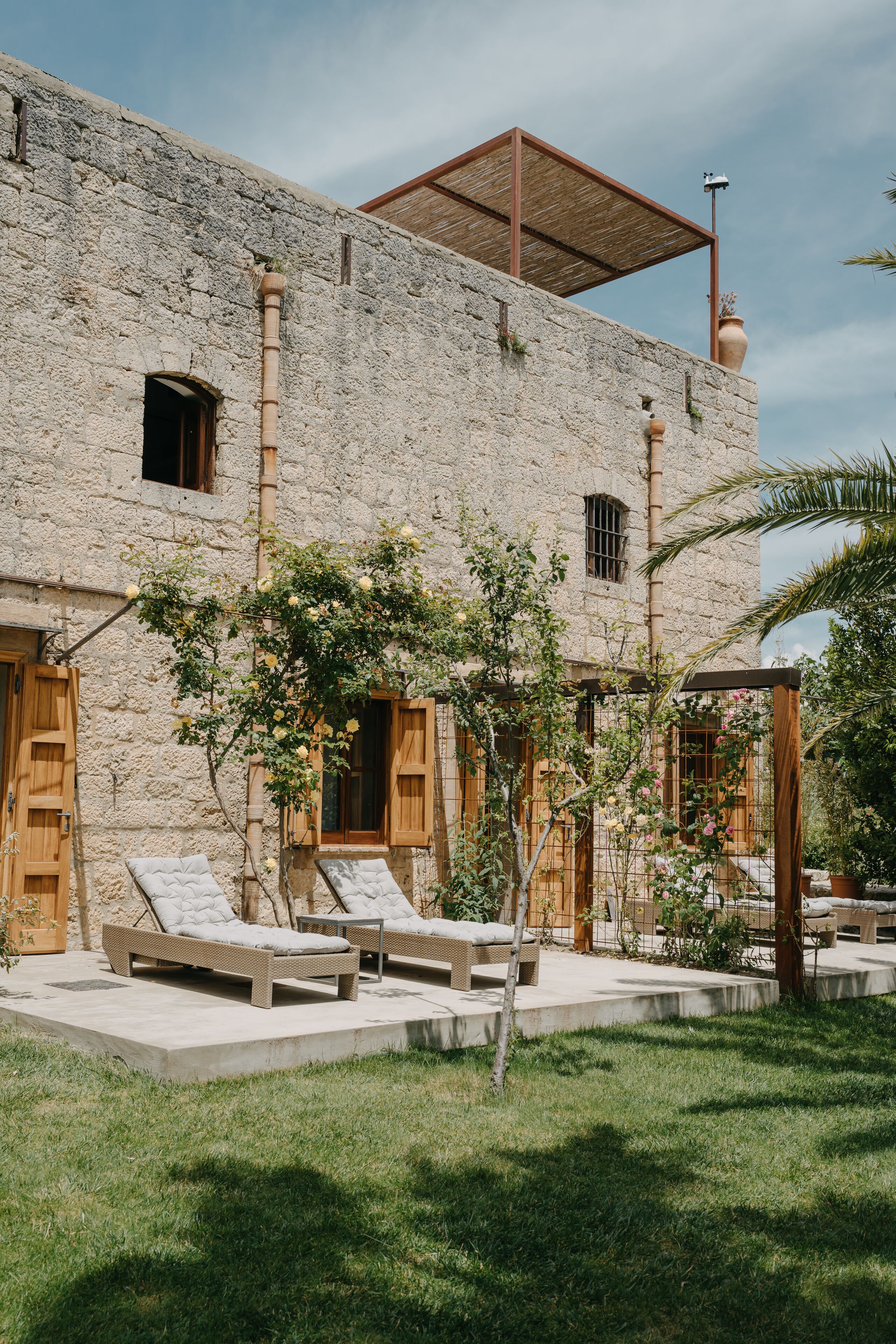 Un edificio de piedra con una pérgola de madera, asientos exteriores y un pequeño jardín.