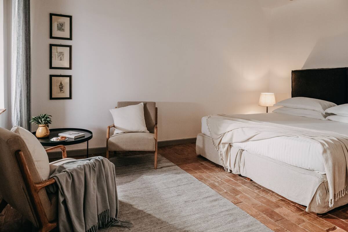 A hotel room with a bed, a chair, a small table with a lamp, and framed pictures on the wall.