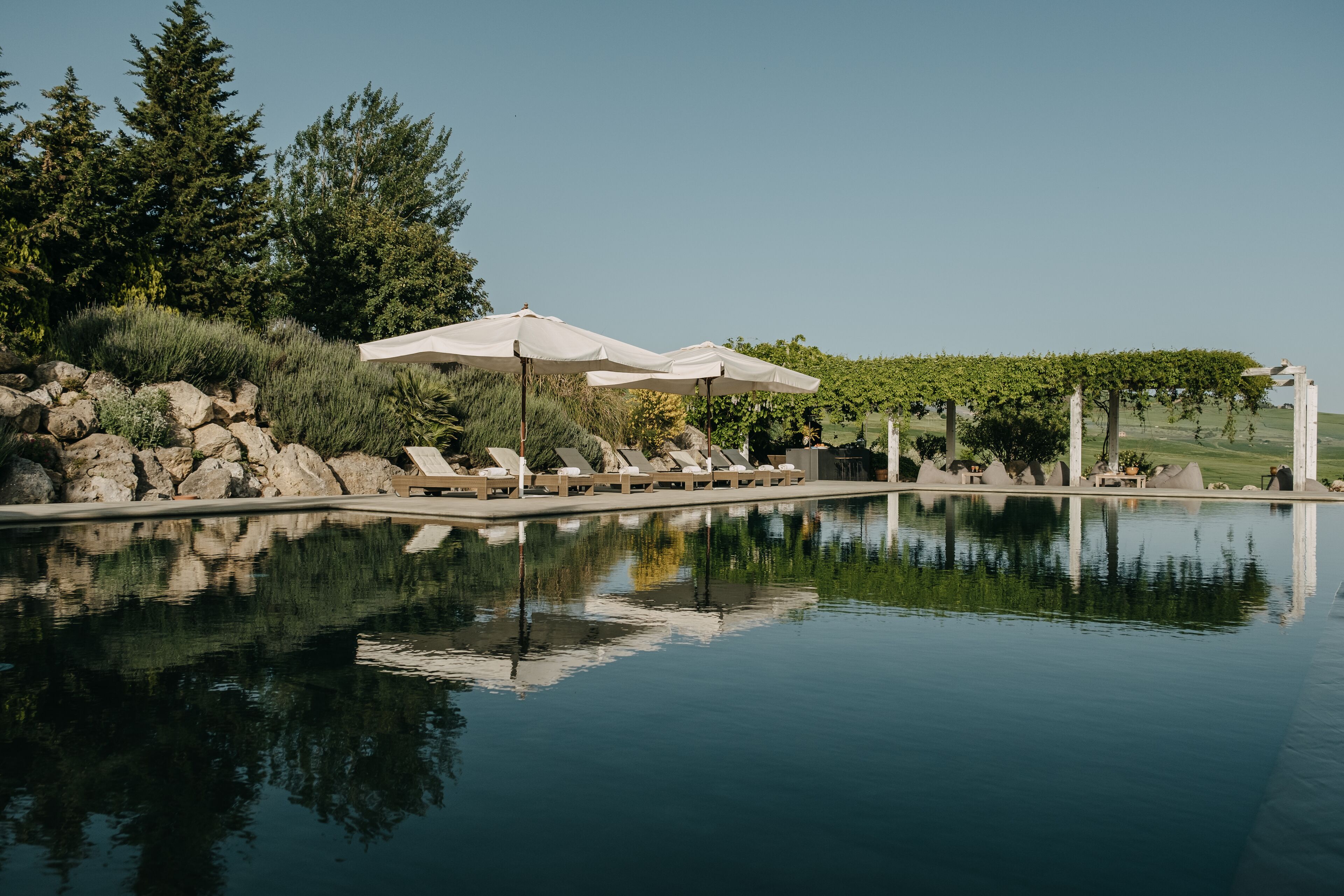 A serene pool area with lounge chairs and umbrellas, surrounded by lush greenery and a clear sky.