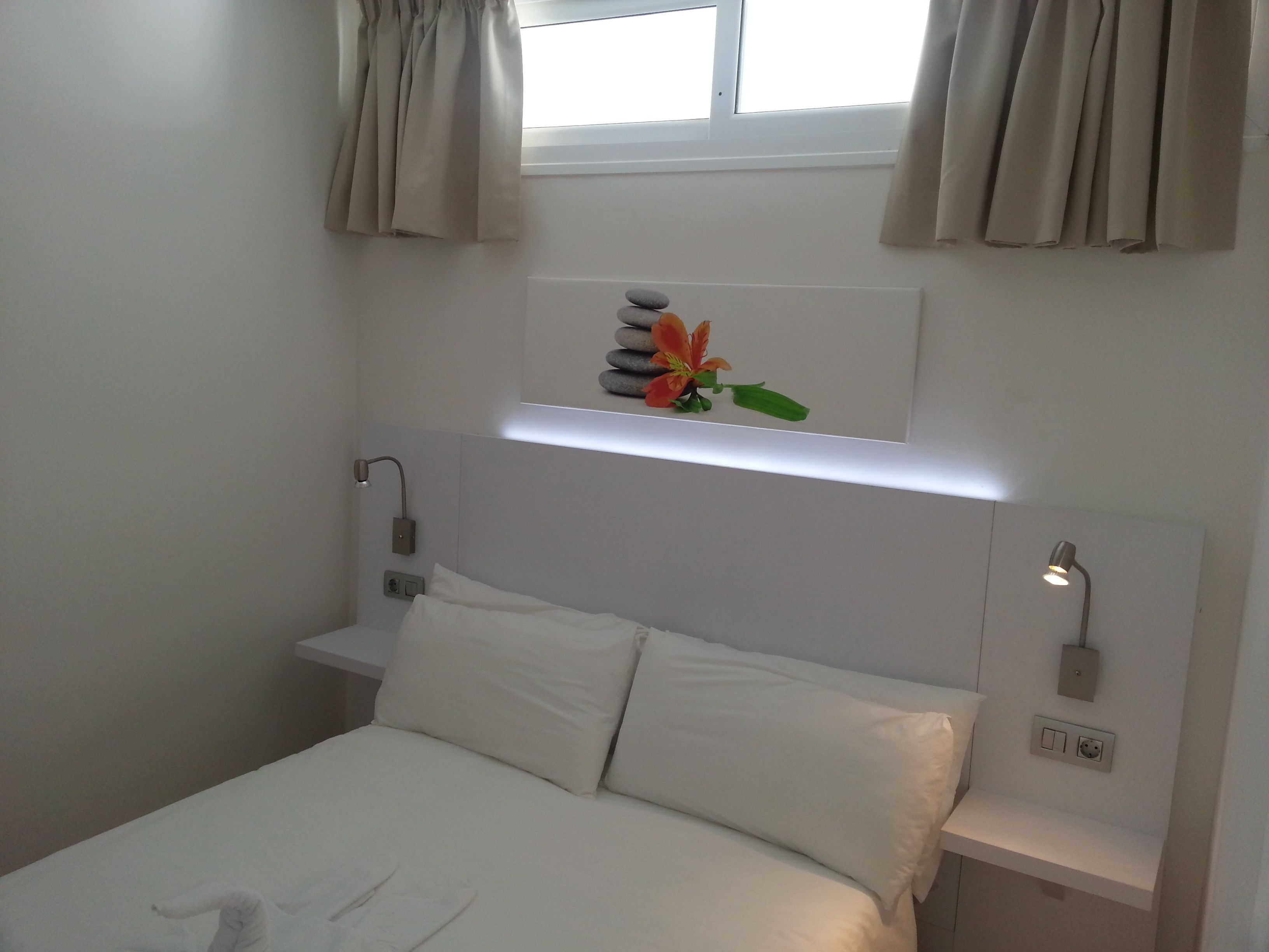 Ein ordentlich eingerichtetes Hotelzimmer mit Bett, Nachttisch und einer Wandablage mit Dekosteinen und einer Blume.