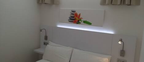 Une chambre d’hôtel bien rangée, avec un lit, une table de chevet et une étagère fixée au mur, ornée de pierres décoratives et d’une fleur.