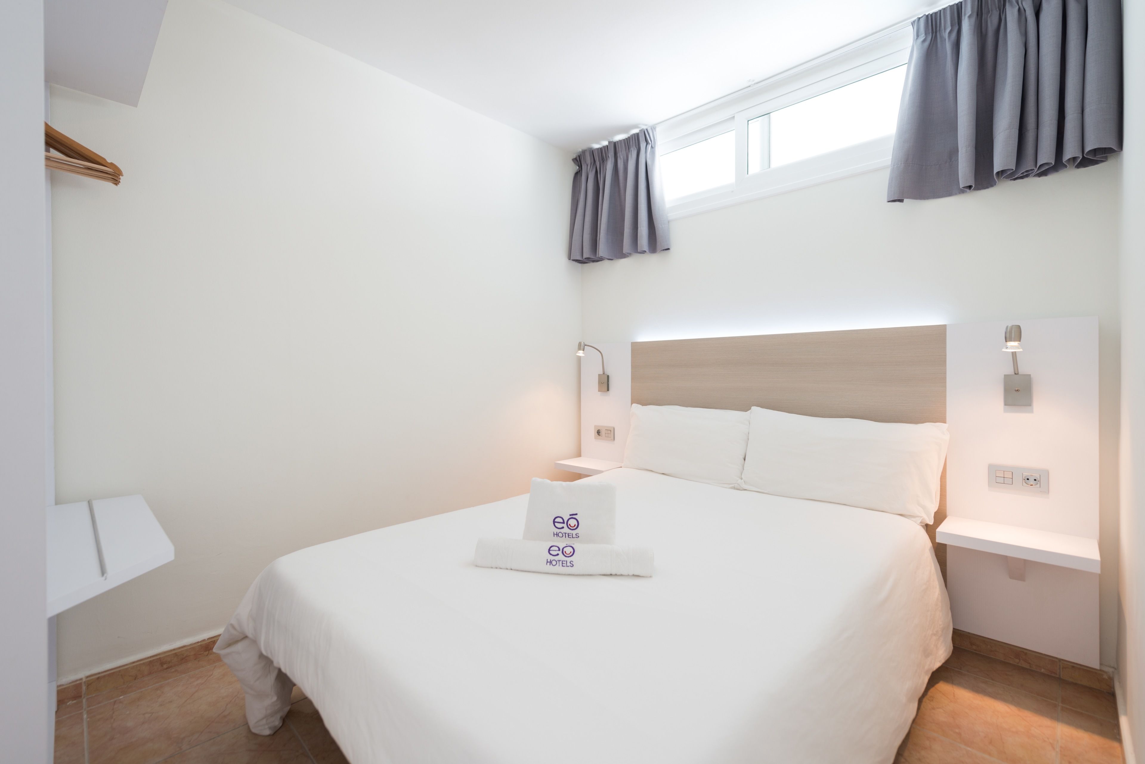 A hotel room with a neatly made bed, a bedside table with a lamp, and a window with curtains.