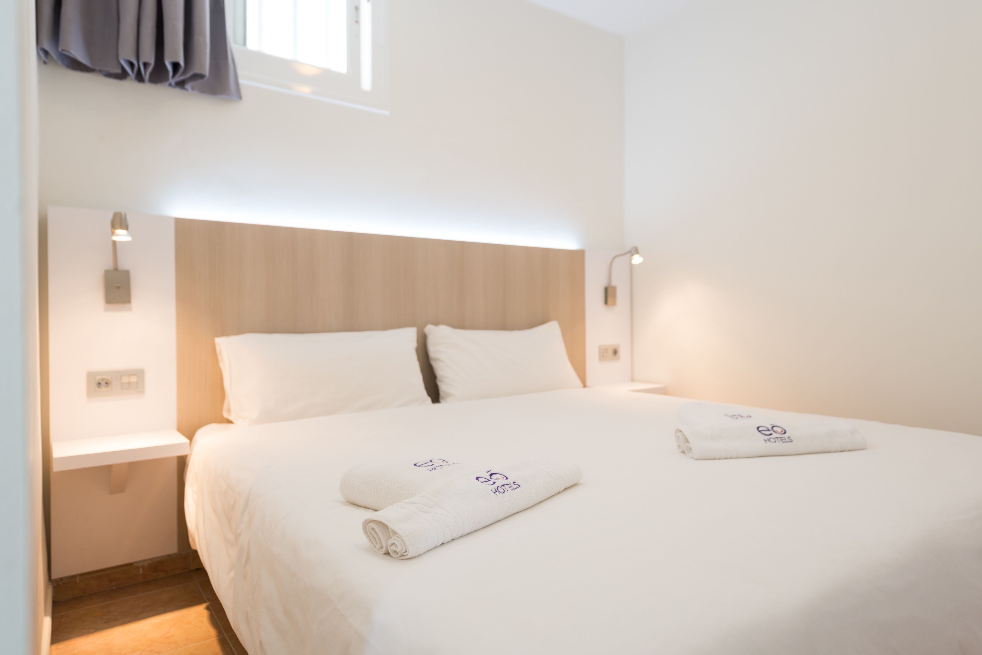 Una cama bien hecha con sábanas y toallas blancas, un cabecero de madera y lámparas de noche.