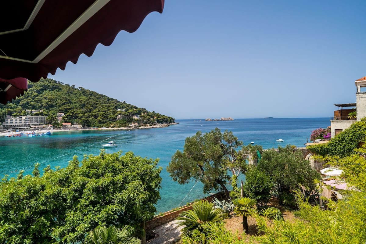A coastal view with a clear blue sea, a hillside with trees, and a building with a balcony overlooking the water.