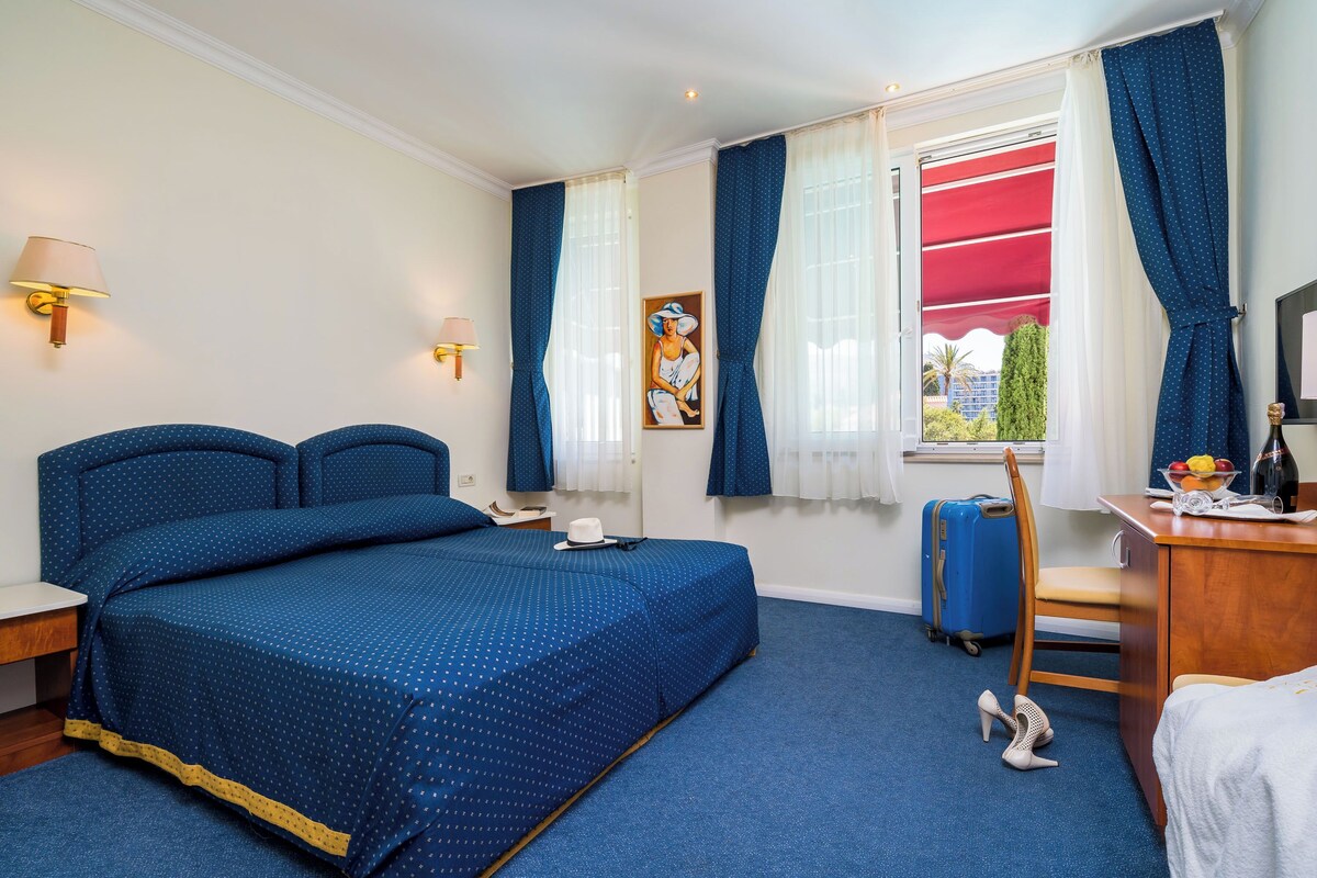 A hotel room with a large bed, a desk, a chair, a suitcase, and a window with curtains.