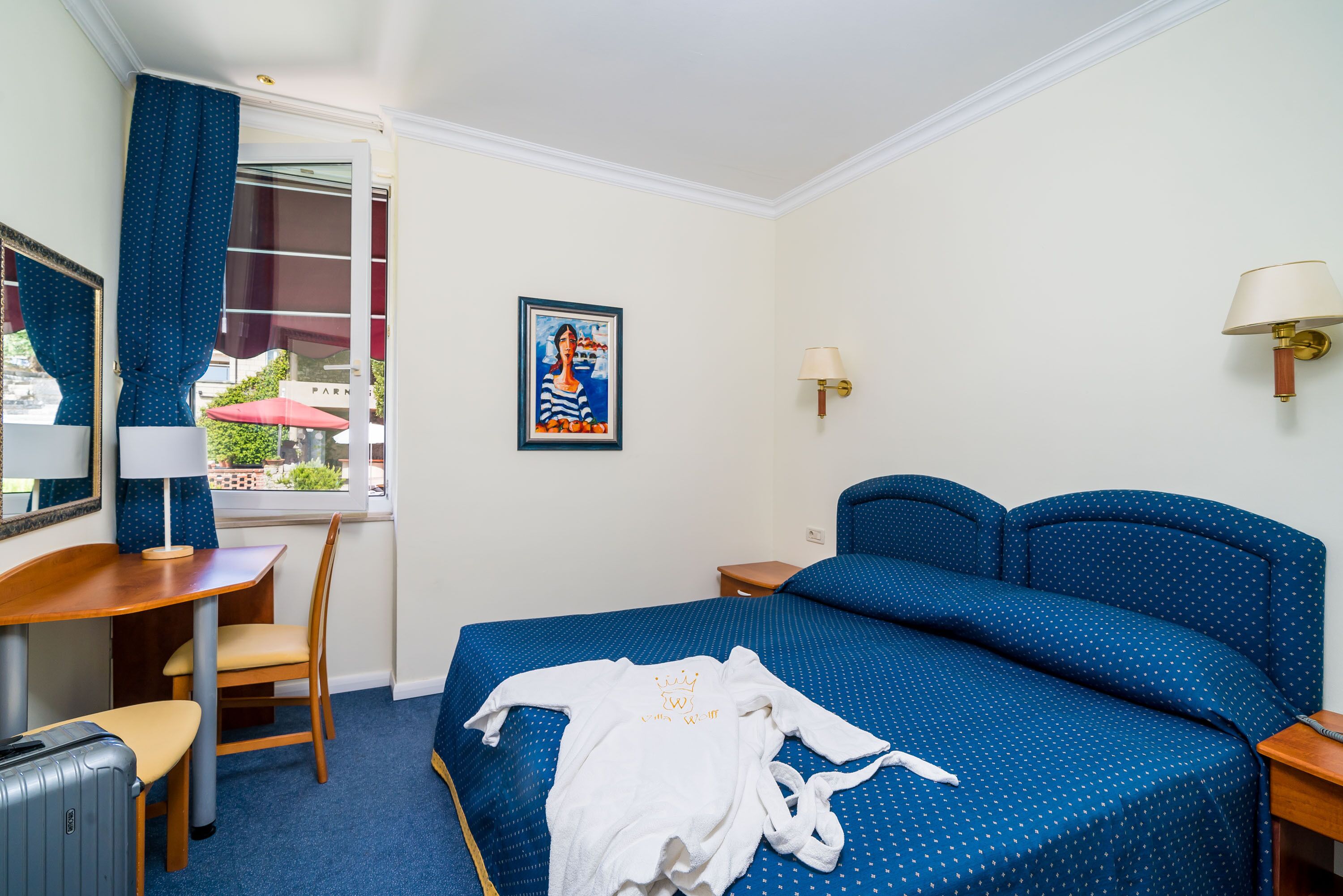A hotel room with a bed, a desk, a chair, a suitcase, and a window with curtains.