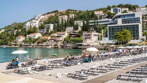 A beach with rows of sun loungers and umbrellas, a rocky shoreline, and buildings in the background.