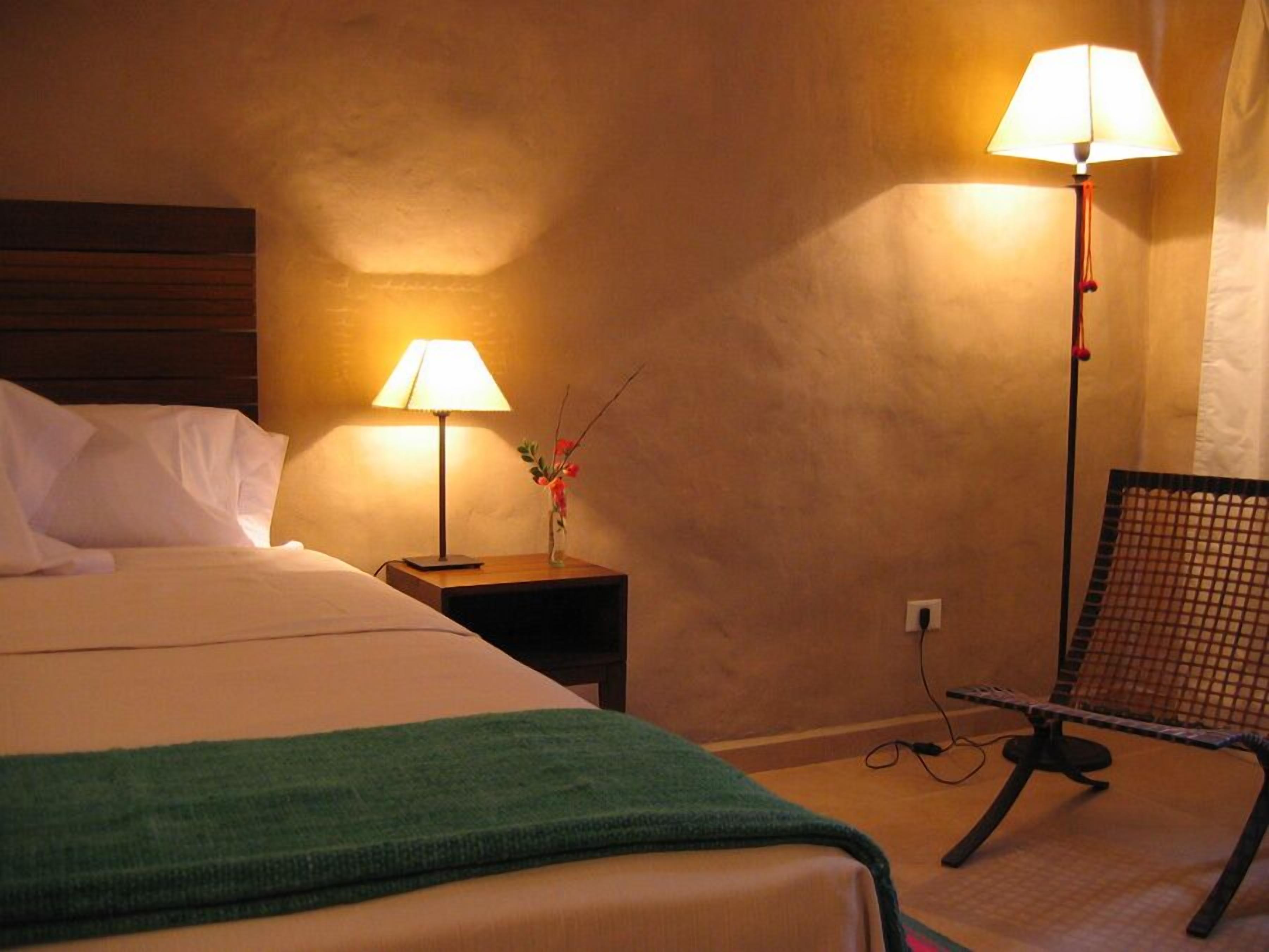 A hotel room with a bed, two bedside lamps, a small table with a vase, and a chair.