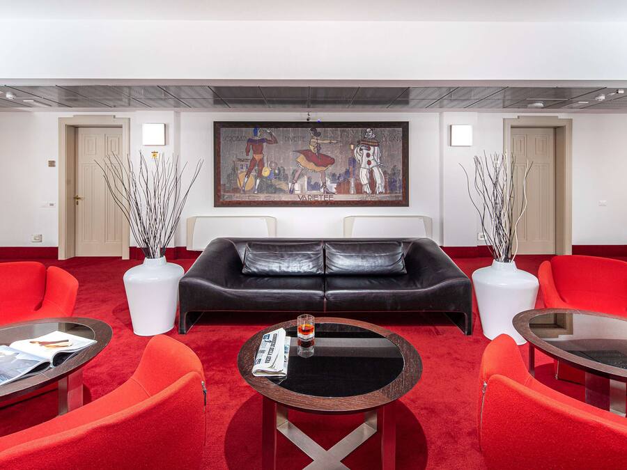 A modern hotel lobby with a red carpet, a black leather sofa, red chairs, and a round table with a magazine.