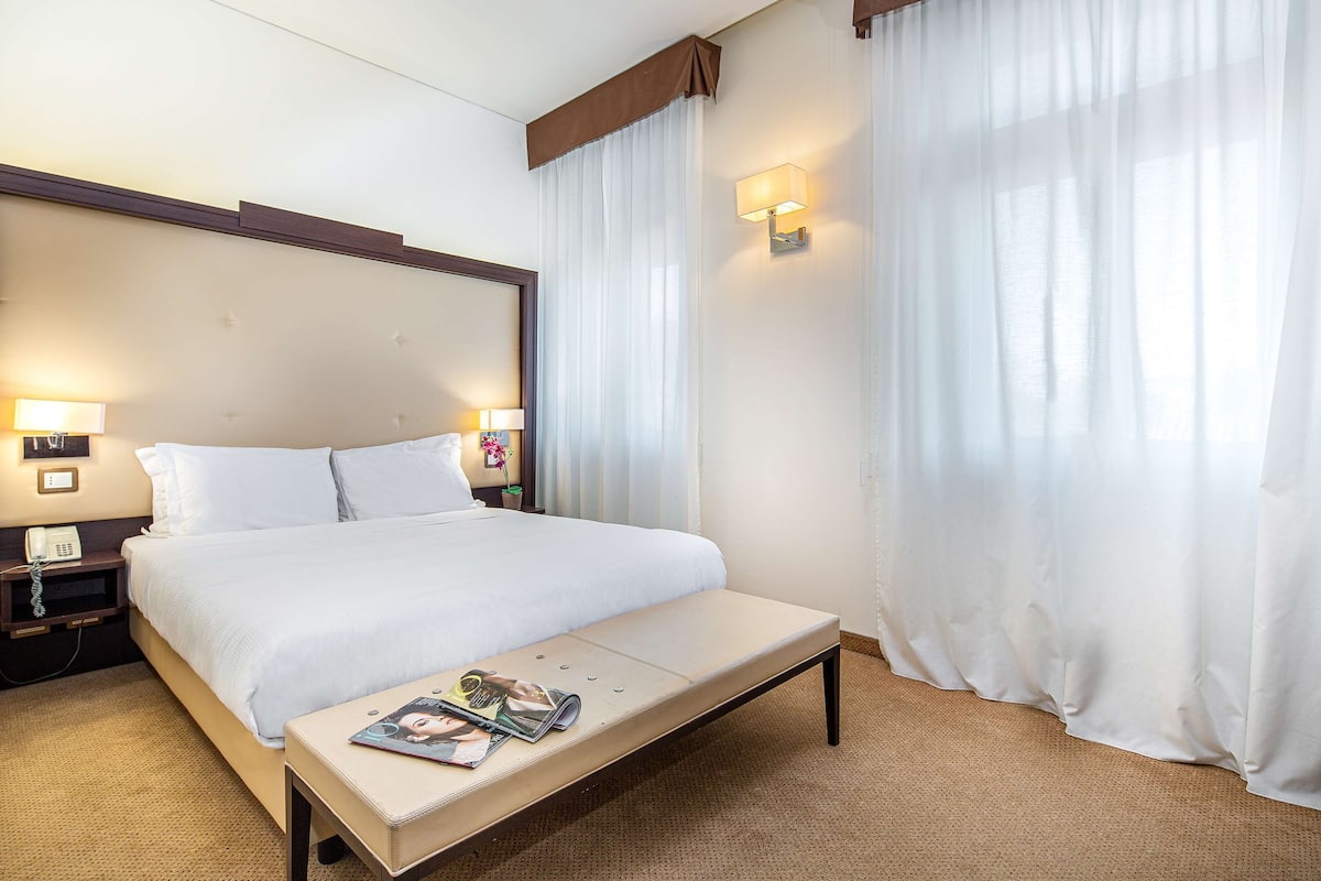 A hotel room with a large bed, bedside lamps, a bedside phone, a magazine on the nightstand, and sheer curtains.