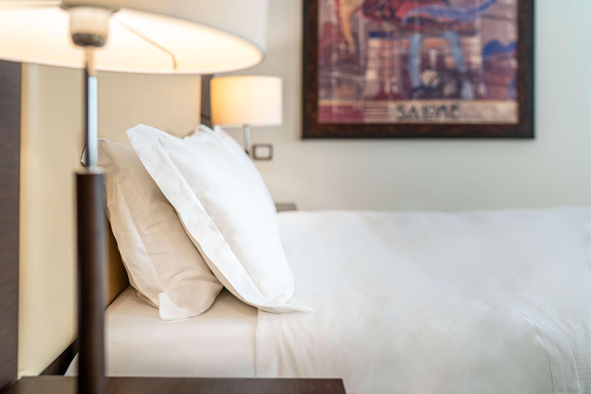 A neatly made bed with white bedding and pillows, a bedside lamp, and a framed picture on the wall.