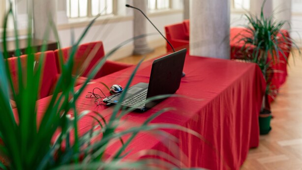 Ein Zimmer mit einem roten Tisch, einem Laptop und einem Mikrofon. Es gibt rote Stühle und Kübelpflanzen.