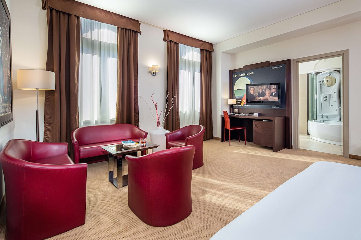A hotel room with a large bed, a TV, a desk, and a seating area with red chairs.