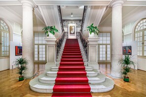 Un magnifique escalier recouvert d’un tapis rouge, bordé de colonnes et agrémenté de plantes en pot, menant à une porte sur laquelle est accrochée une image encadrée.