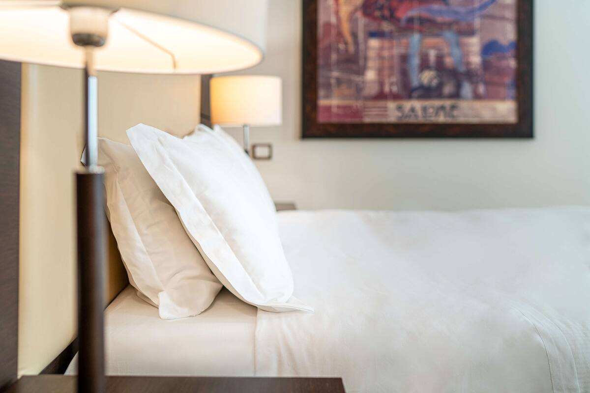A neatly made bed with white linens and pillows, a bedside lamp, and a framed picture on the wall.
