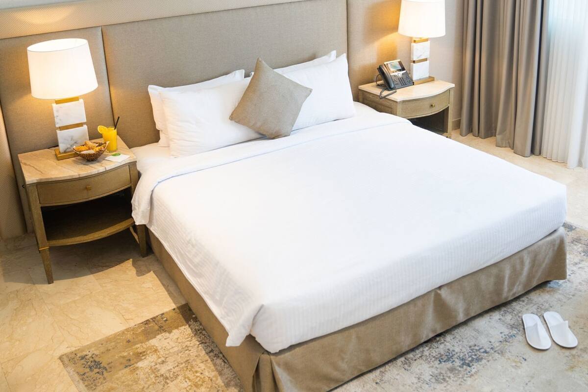 Een hotelkamer met een groot bed, twee nachtkastjes met lampen, een telefoon en een paar slippers op de vloer.