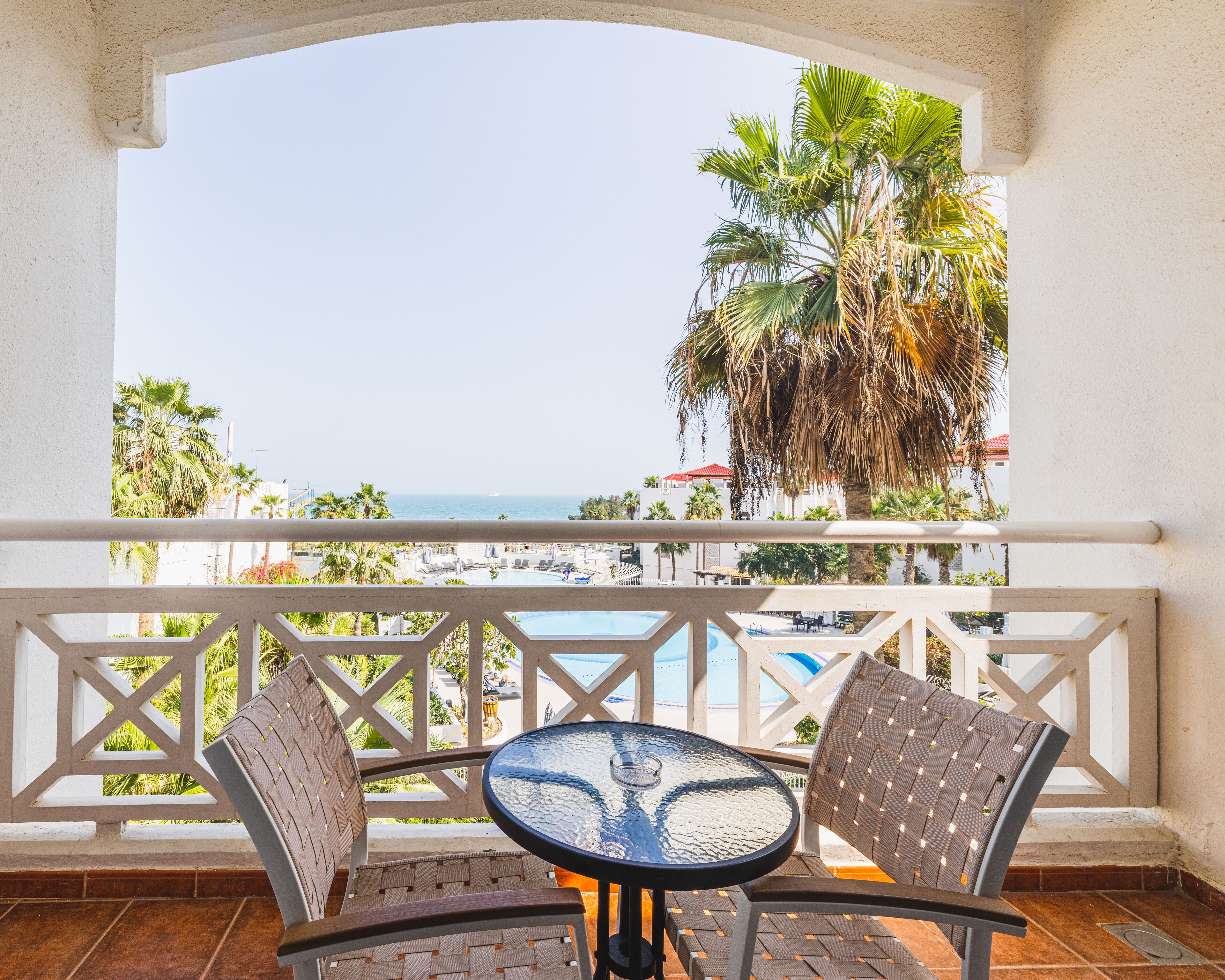 A balcony with a glass table and chairs, overlooking a pool and palm trees.