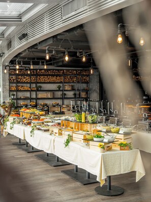 A buffet setup with various food stations, a long table with a white tablecloth, and a modern industrial ceiling with exposed pipes and lighting.