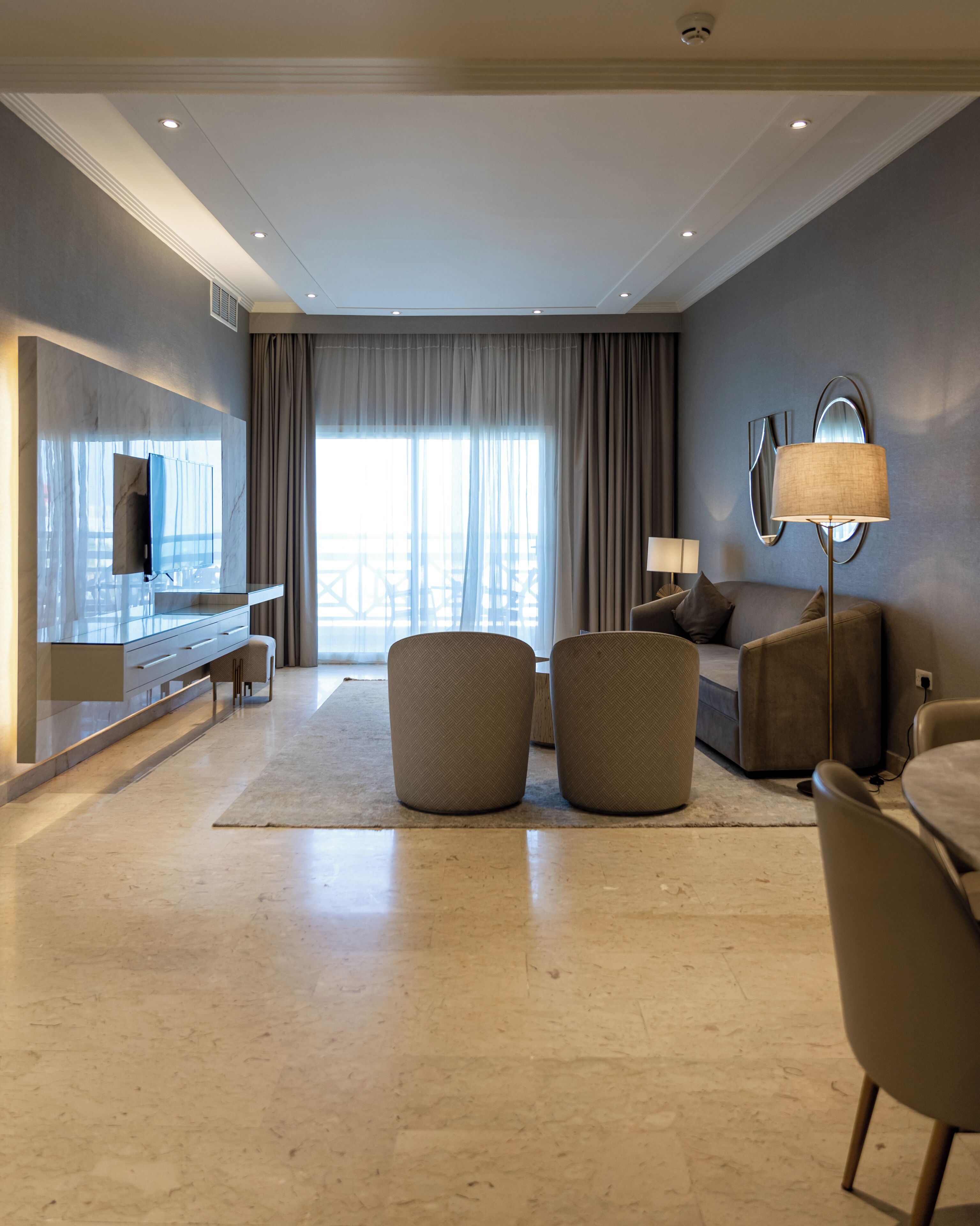 A modern hotel room with a flat-screen TV, a sofa, two ottomans, a lamp, and a large window with curtains.