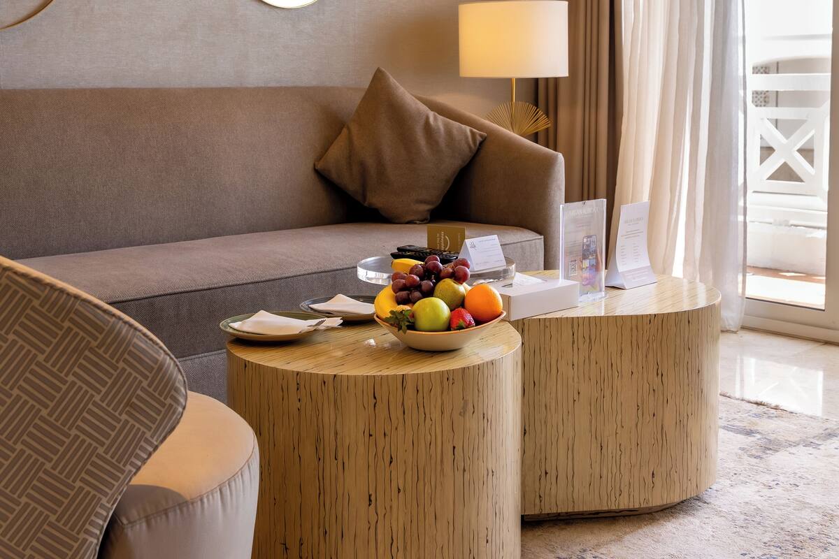 Een moderne hotelkamer met een bank, twee ronde houten salontafels, een fruitschaal, een lamp en een grote spiegel aan de muur.