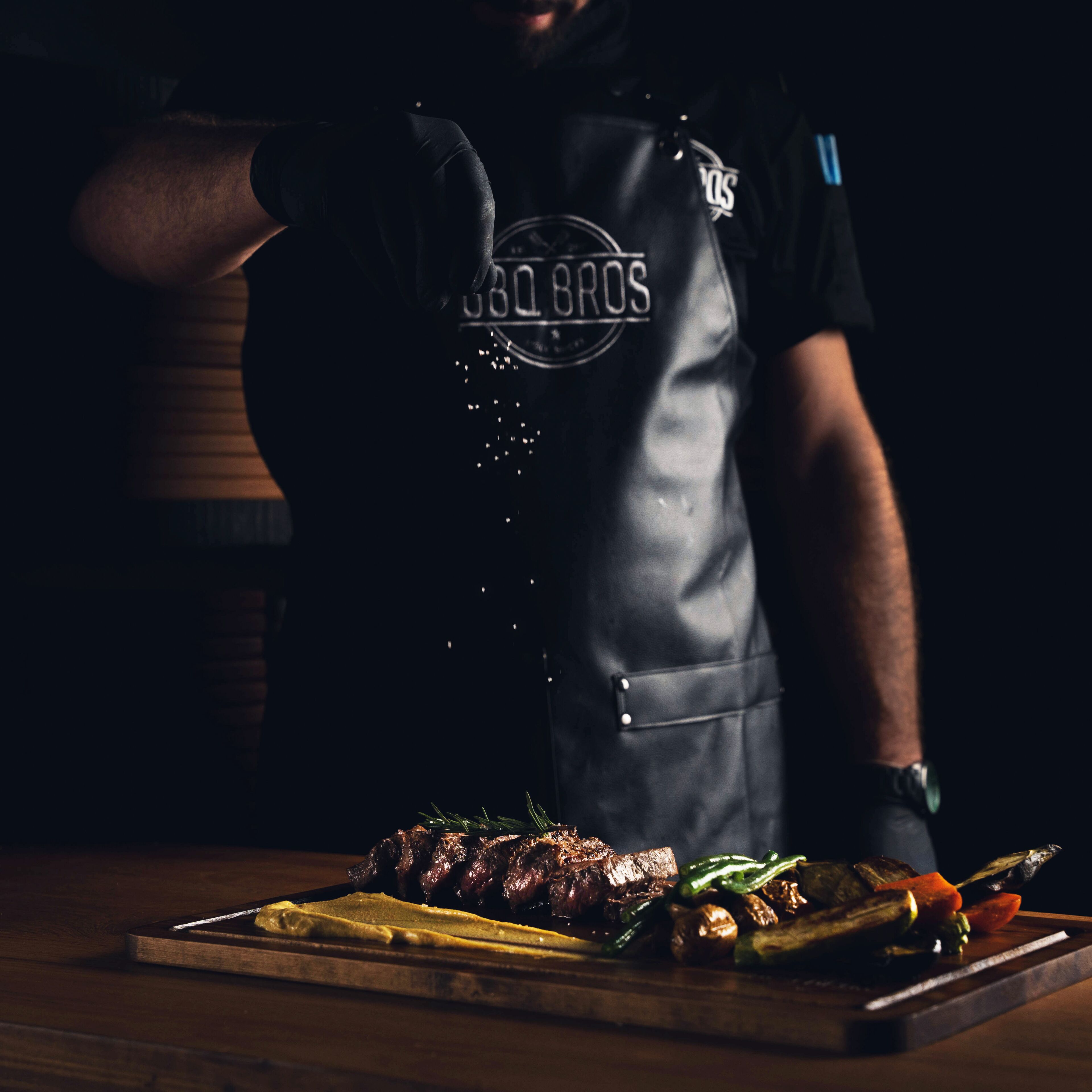 Una persona con delantal negro que lleva la inscripción "BBQ BROS", de pie detrás de una mesa de madera con un plato de comida que incluye carne y verduras.