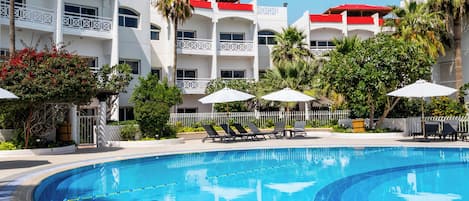 Una piscina con borde de azulejos, rodeada de sillones de descanso y sombrillas, con un edificio con balcones al fondo.