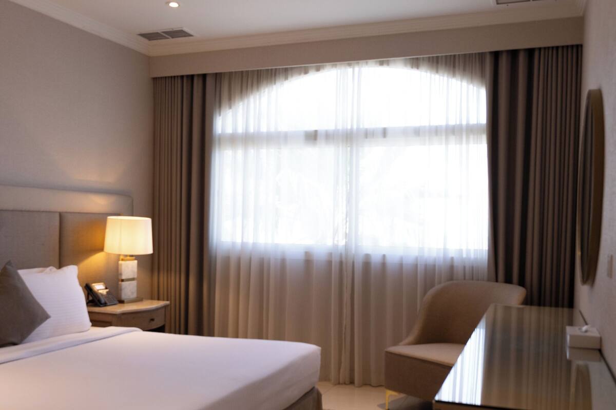 Hotelkamer met een bed, bureau, stoel, lamp, raam met gordijnen en een aan de muur bevestigde televisie.