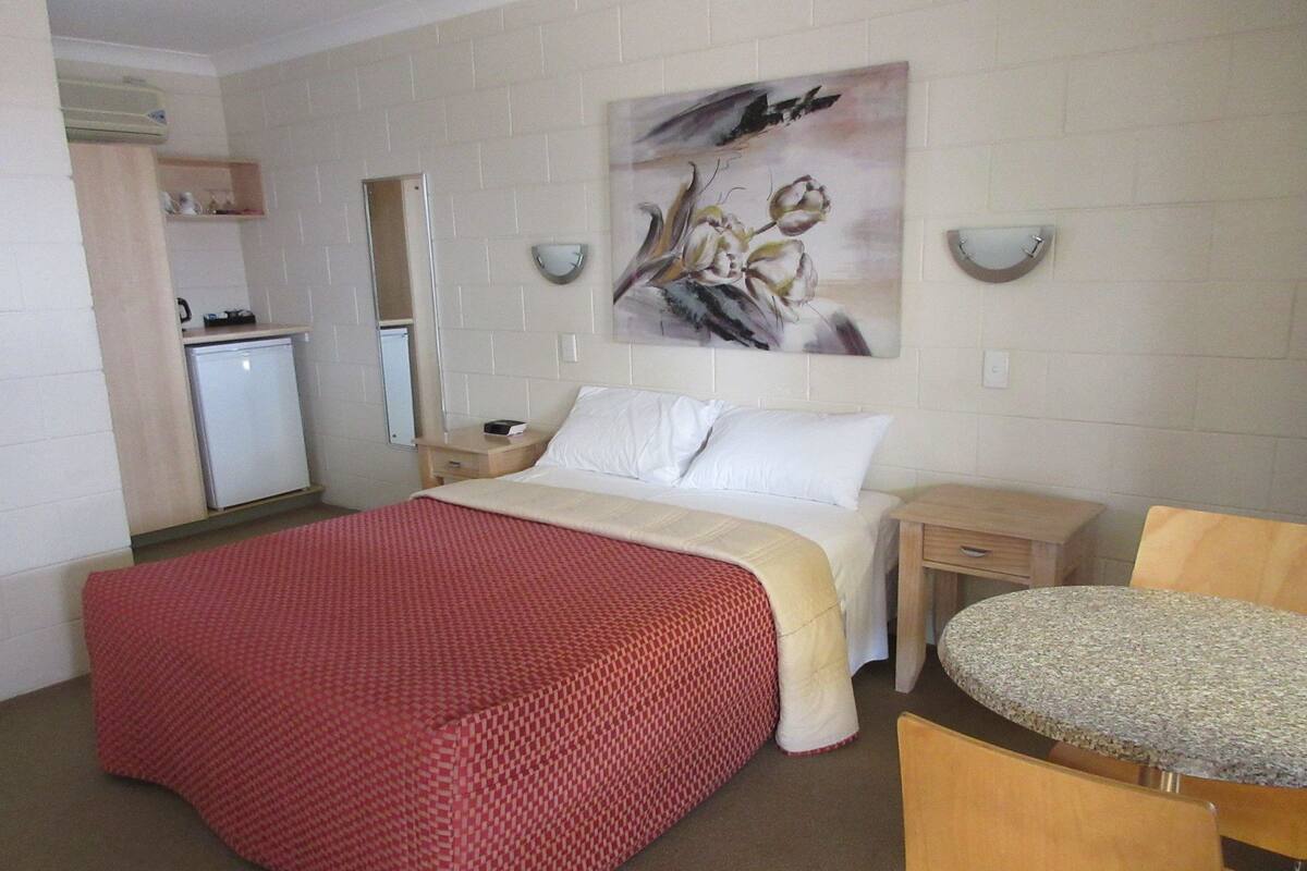 Una habitación de hotel con cama, escritorio, silla, kitchenette pequeño y un cuadro en la pared.