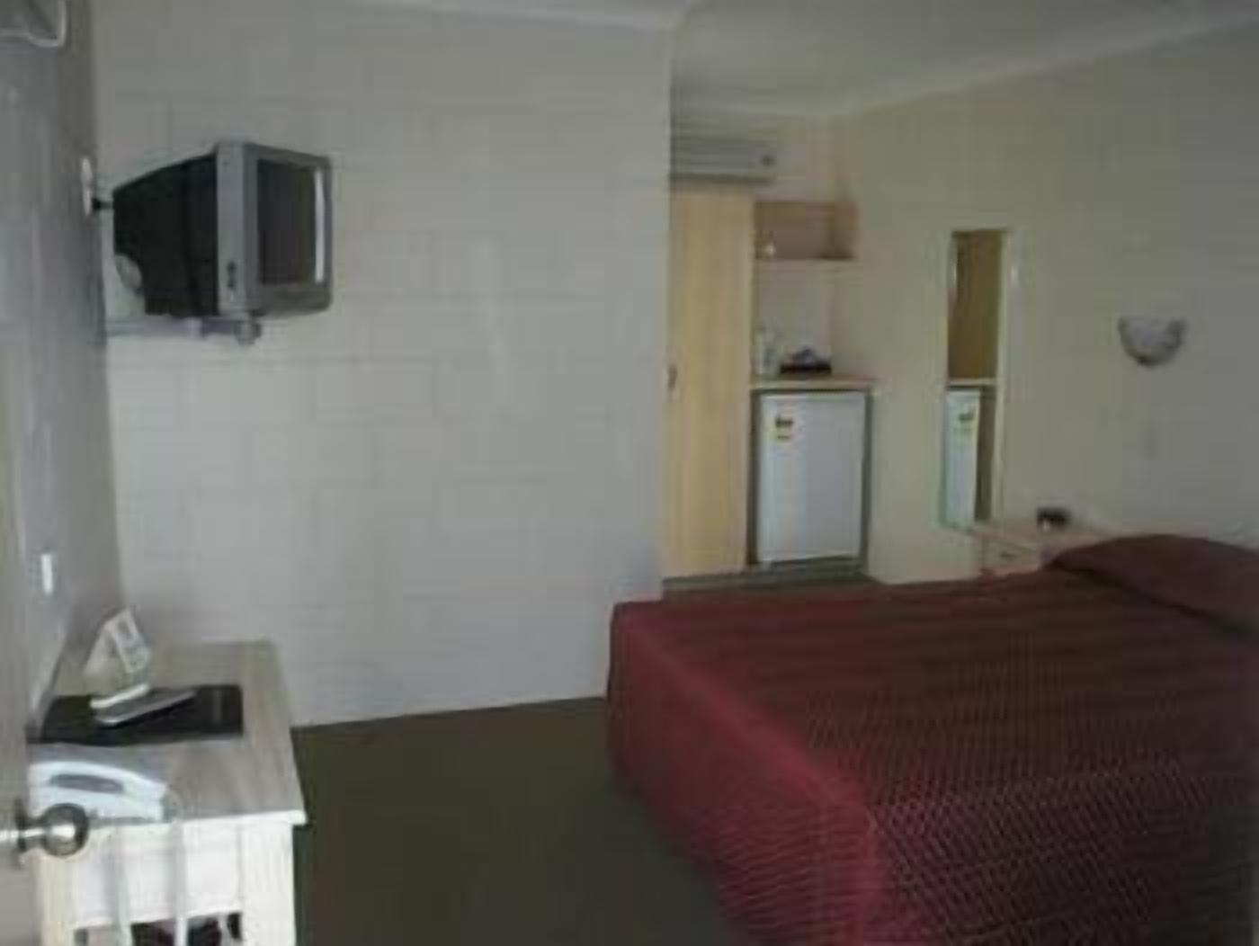 A hotel room with a bed, a desk with a computer, a television mounted on the wall, and a small kitchenette.