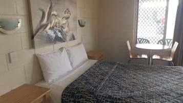 A hotel room with a bed, a nightstand, a painting on the wall, a window with a view, and a table with chairs.