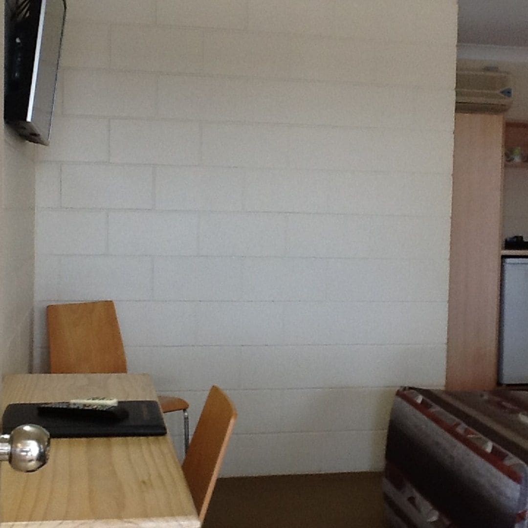 Una habitación pequeña y poco amoblada, con una mesa y sillas de madera, un televisor en la pared y una alfombra estampada en el suelo.