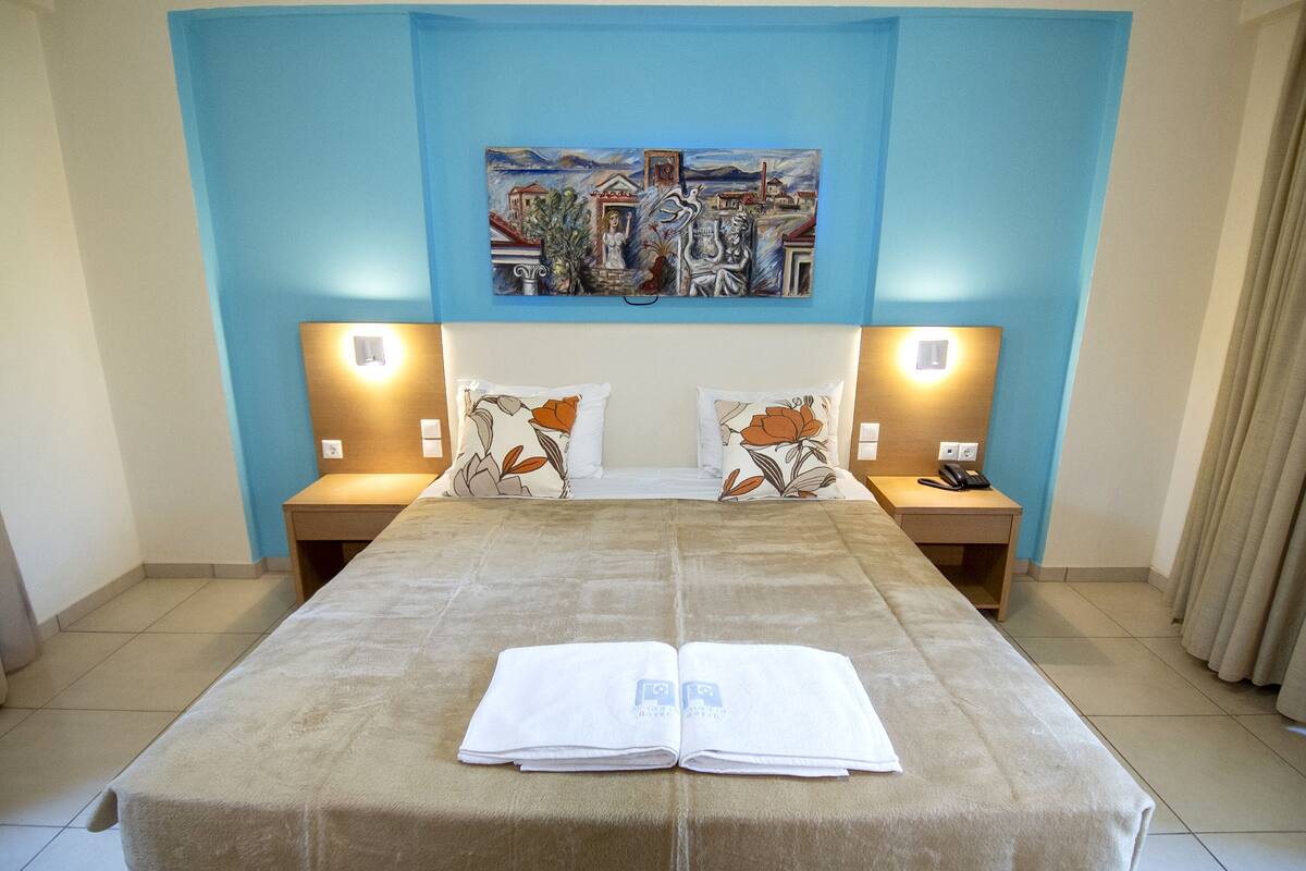 A hotel room with a large bed, two bedside tables, a painting on the wall, and a window with curtains.