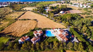 Vista aérea de un complejo turístico costero con varios edificios, una gran piscina y campos circundantes.