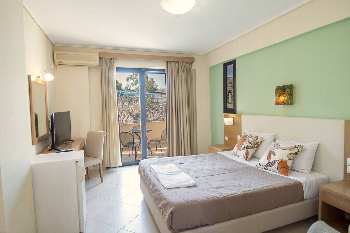 A hotel room with a bed, a desk, a chair, a TV, and a balcony with a view.