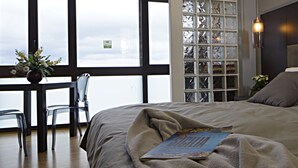 A bedroom with a large bed, a bedside table with a vase of flowers, a chair, and a view of the city through a window with a decorative glass panel. - Venere B&B (Catania)