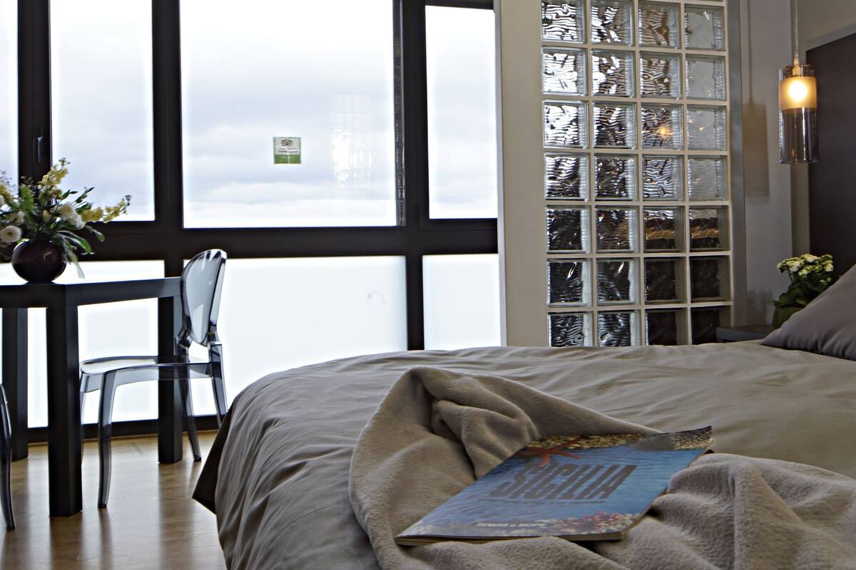 A bedroom with a large bed, a bedside table with a vase of flowers, a chair, and a view of the city through a window with a decorative glass panel.