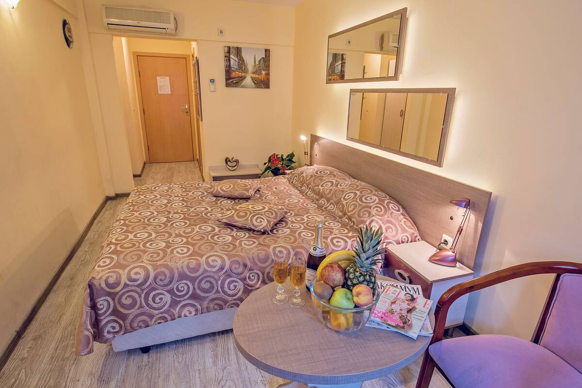 Habitación de hotel con cama, mesita de noche con lámpara, una silla, una pequeña mesa con bebidas y fruta, y un espejo instalado en la pared.