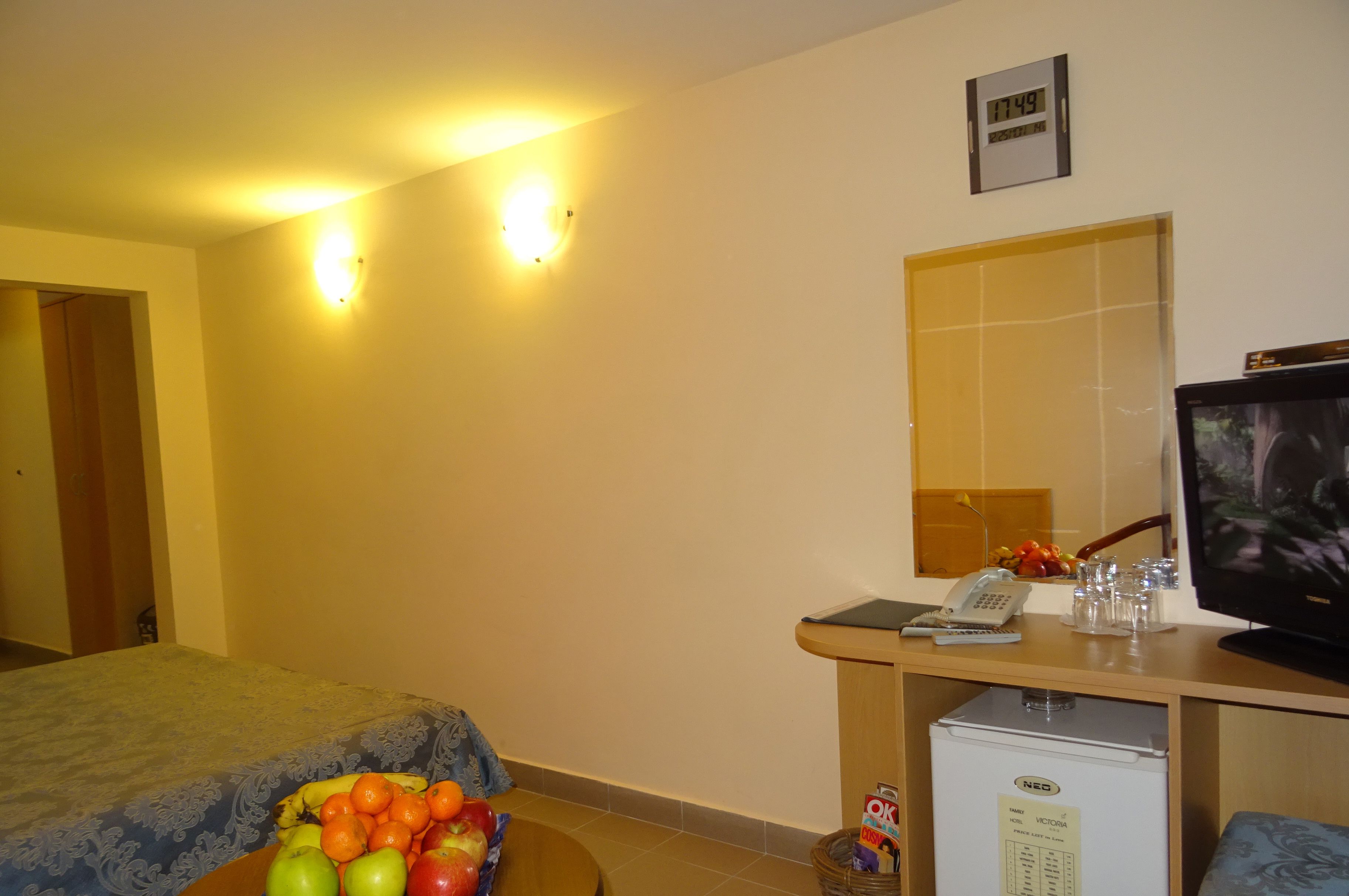 A hotel room with a bed, a small desk with a TV, a refrigerator, and a fruit bowl.