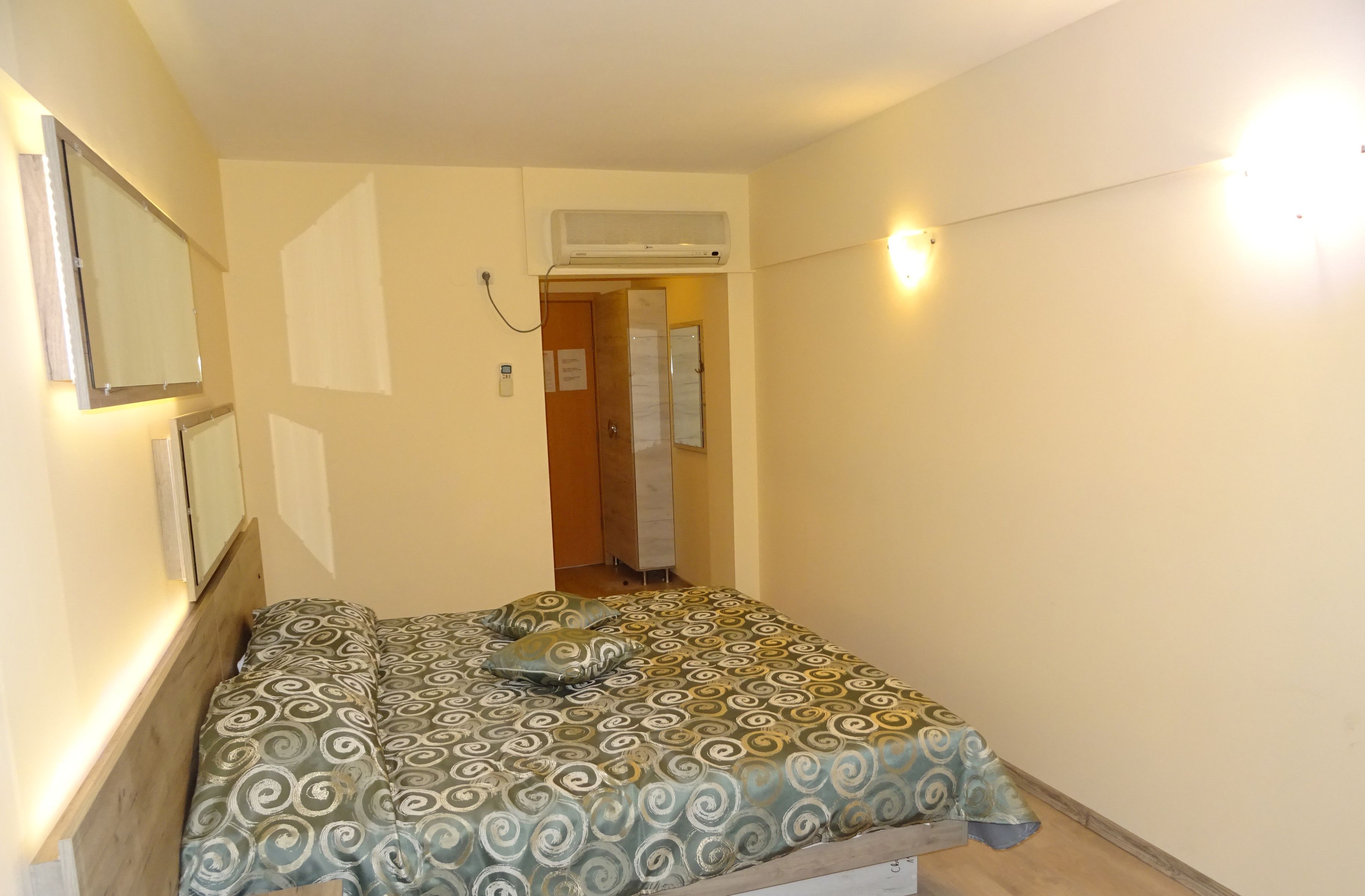 A small, narrow room with a bed, a mirror, and an air conditioning unit.