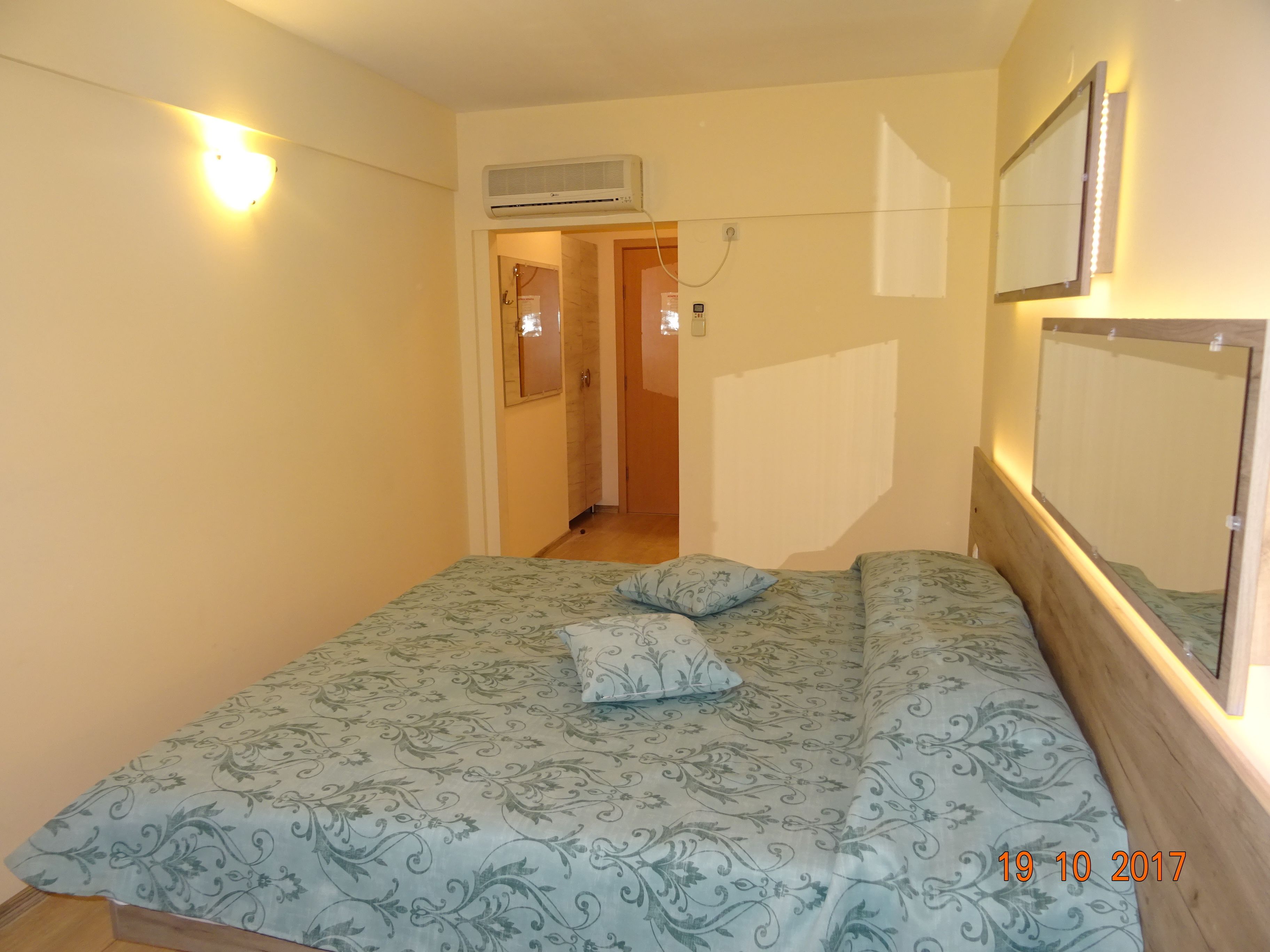 A bedroom with a bed, two pillows, an air conditioning unit, a mirror, and a door leading to another room.
